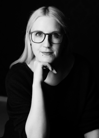 Portrait photo of Dana. A person presenting as a woman with bold black glasses, blonde long hair and facial piercings. They wear a black jumper and smile at the camera.